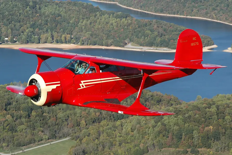 A vibrant red vintage airplane flying over a scenic landscape with river and forest.