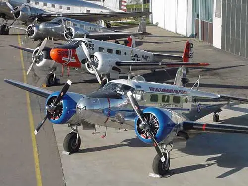 Several vintage airplanes parked on a tarmac near hangars.