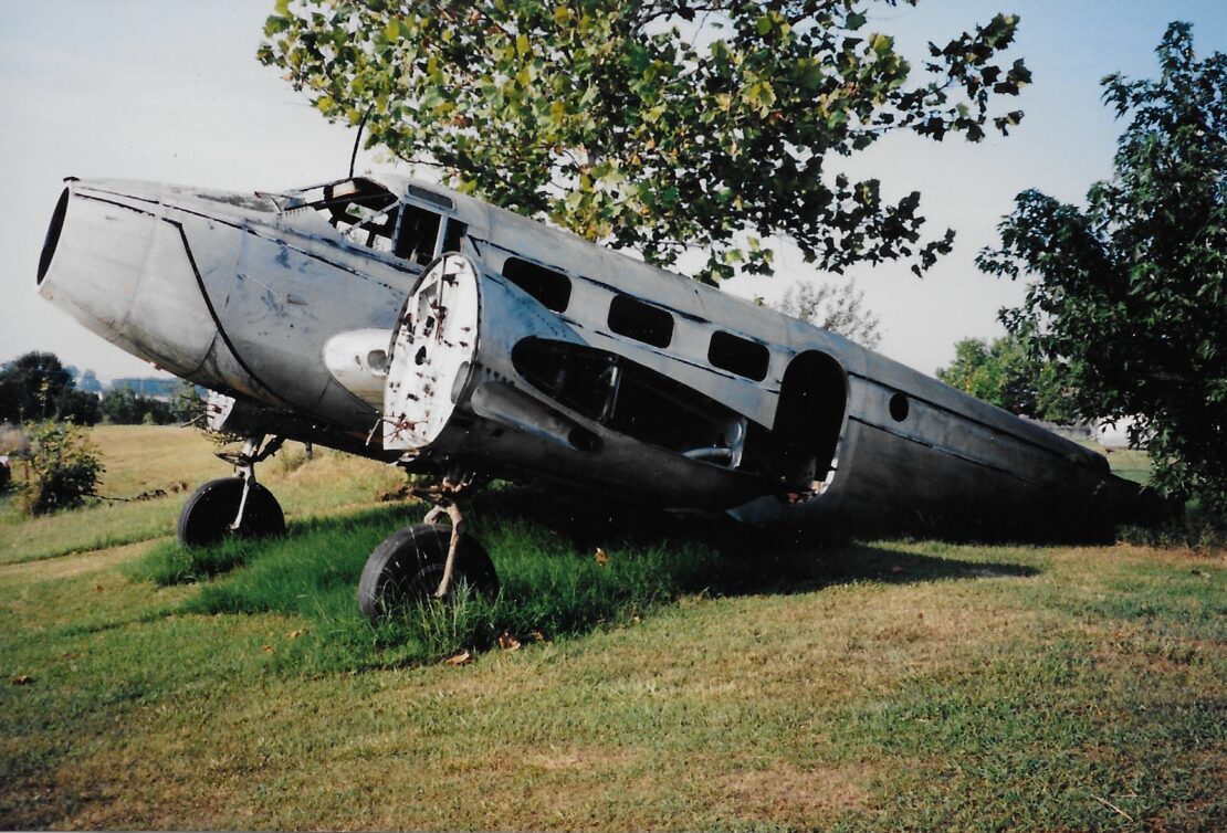 Abandoned, rusty airplane fuselage resting on grass under a tree.
