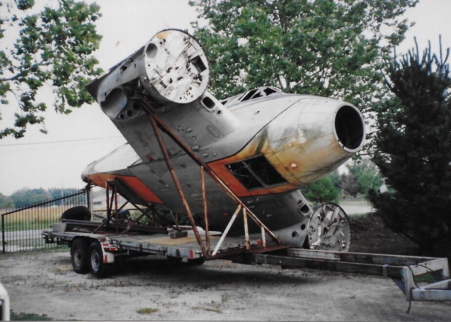 An old, weathered aircraft nose and engines on a trailer outdoors.